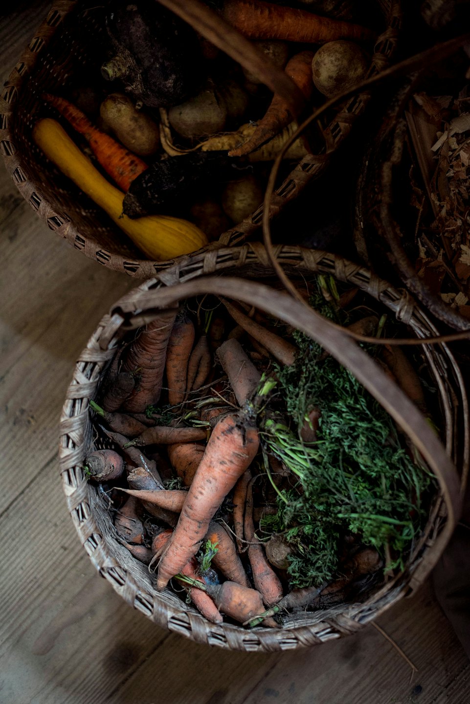 Two wicker baskets full of fresh carrots and squash on a wooden floor.
