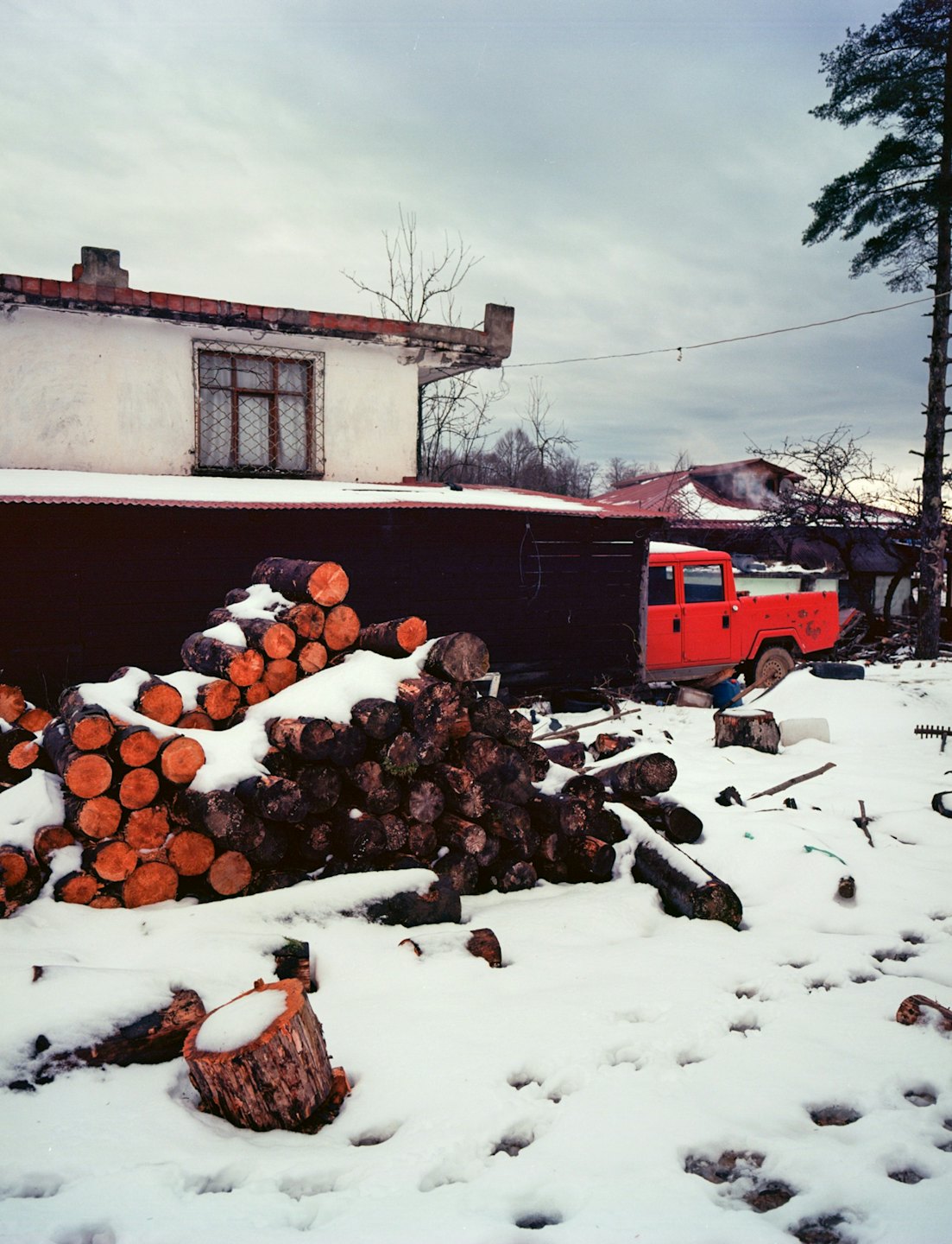 Snow covered firewood stacked outside a rural house and shed.