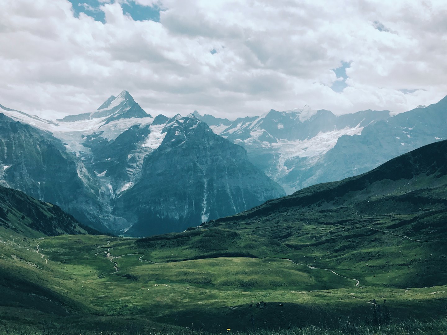 Dirt paths wind through a green valley toward snow-capped mountains.